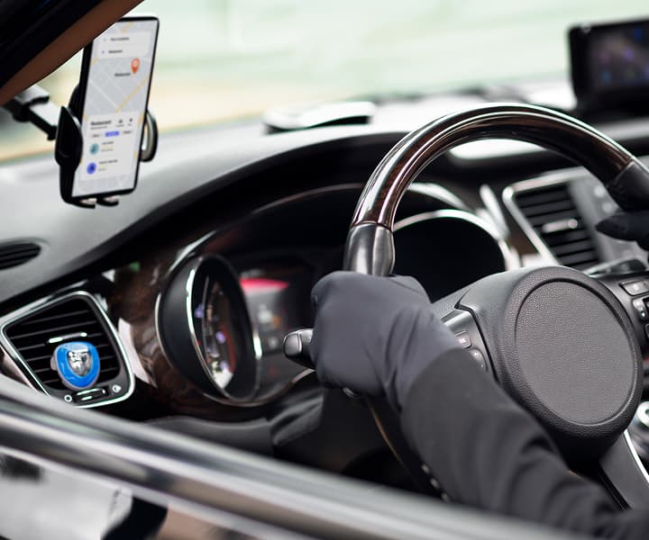 A driver's perspective inside a modern car, showing hands on the steering wheel and a smartphone mounted on the dashboard displaying a GPS navigation map.