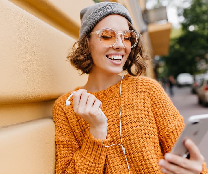 A smiling young woman wearing a yellow knitted sweater, a gray beanie, and orange-tinted glasses, holding a smartphone and using wired white earphones.